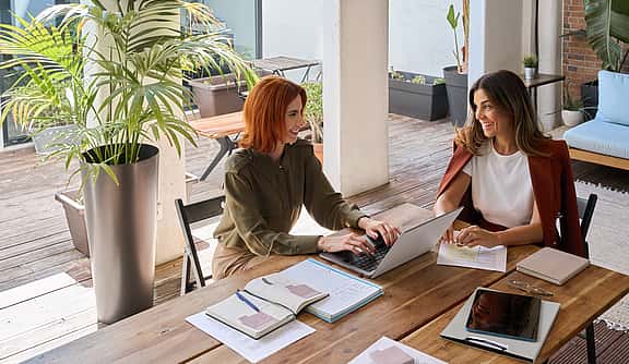 Two happy business women of young and middle age talking in green cozy office sitting at desk