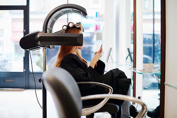 Lady sitting at a hairdresser's chair with rollers in her hair and a professional orbiting hood hair dryer in view, highlighting grenke's financing solutions for beauty equipment, including advanced skincare devices and professional salon furniture to enhance client experiences.