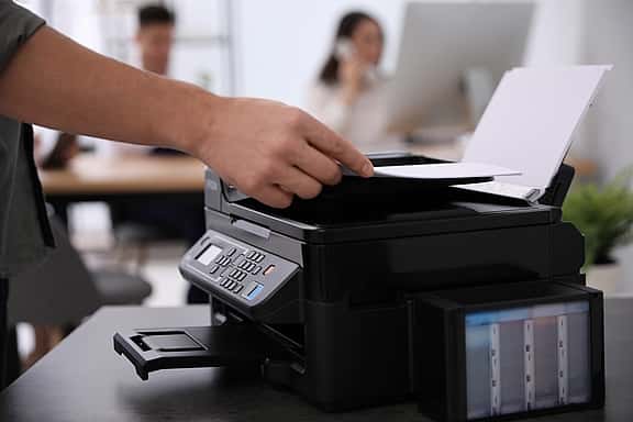 Close-up of hands using a printer in an office, with people working in the background, highlighting grenke's leasing options for laser, LED, multifunction, and 3D printers.