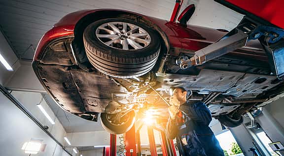 Low-angle view of a car lifted in a garage, with a mechanic inspecting underneath, showcasing grenke's financing solutions for automotive service center equipment including lifts, diagnostic tools, tire changers, and alignment machines.