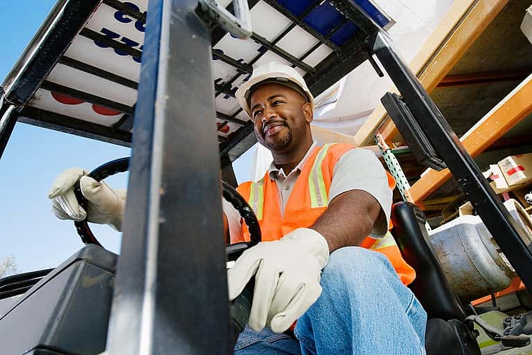 A smiling man in a neon orange vest driving a forklift, illustrating grenke's leasing options for various forklifts and material handling equipment, backed by strong UK partnerships and flexible terms.