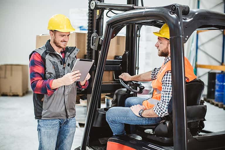 Two men in hard hats discussing work in a warehouse with boxes and shelves in the background, representing grenke's tailored forklift fleet financing solutions for the UK manufacturing and handling industry.