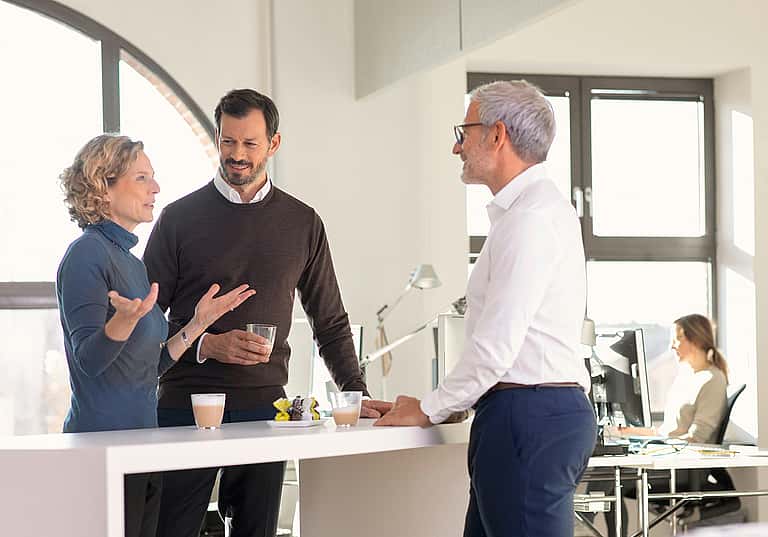Three business people standing in an office, engaged in a conversation about equipment descriptions, maintenance requirements, and grenke's hire agreement terms and conditions.