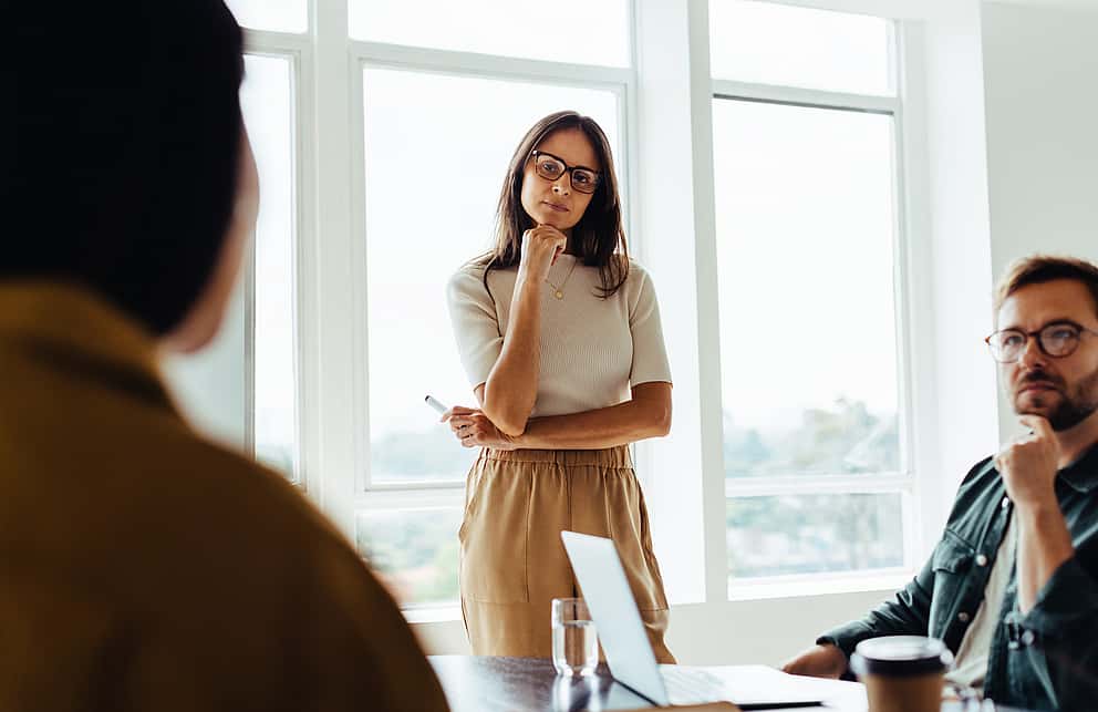 A businesswoman sitting at her desk using her phone, in a busy office environment, symbolizing grenke's commitment to helping ambitious partners grow by offering leasing solutions for sales without boundaries.